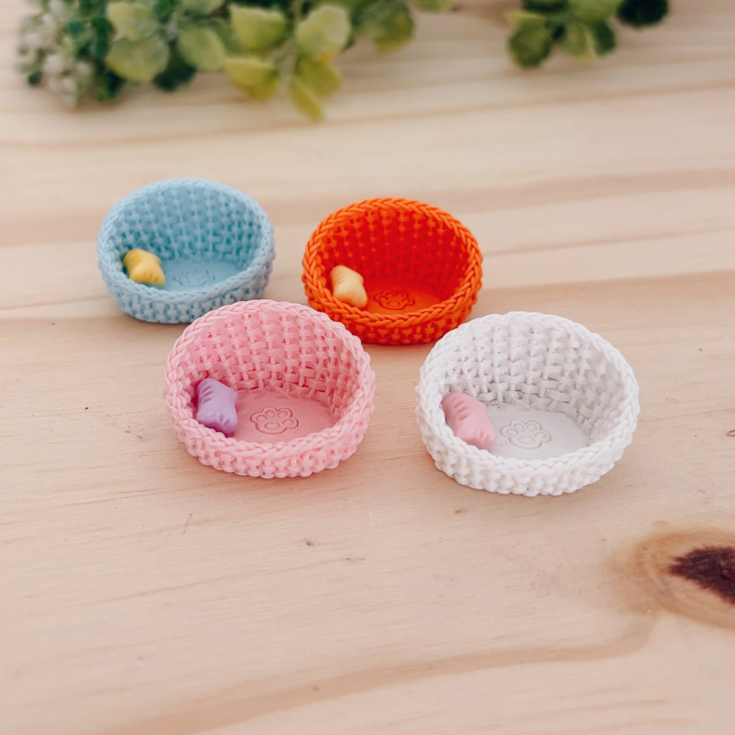 Four miniature wicker pet beds in blue, pink, orange, and white on a wooden surface with greenery in the background.